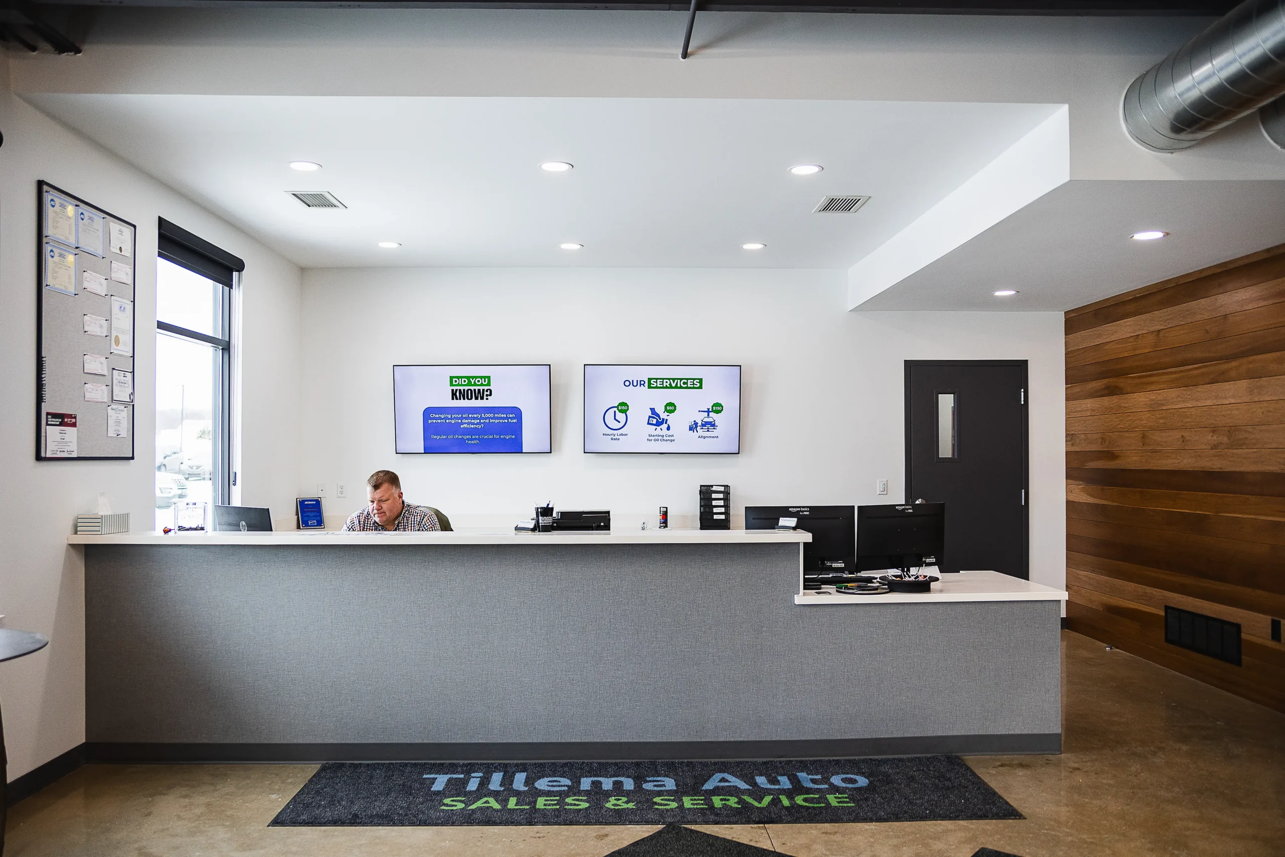 Reception desk at Tillema Auto with a staff member at the counter and digital displays showing company information on the wall behind him.